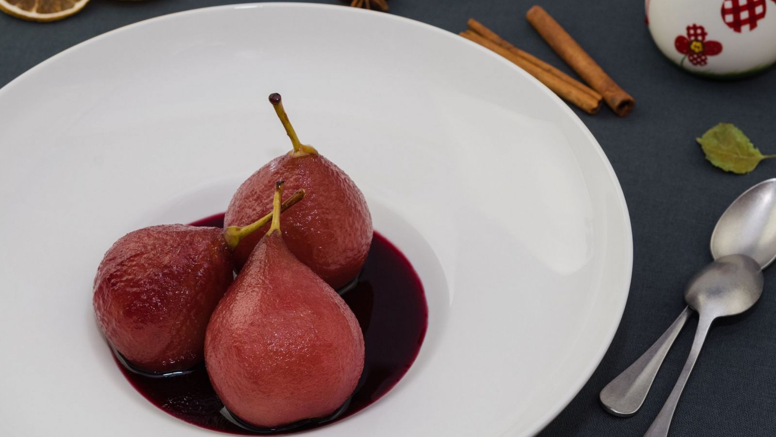 Plato de peras rojas al vino con canela, naranja deshidratada y anís estrellado.  Cucharas y hoja seca completan la escena otoñal.