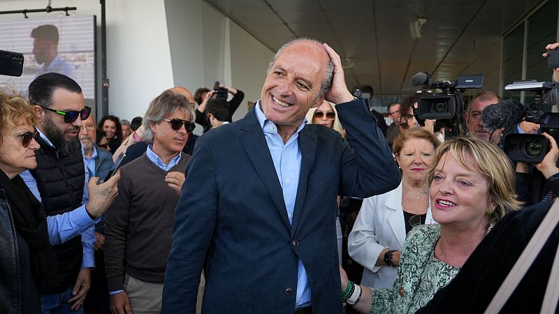 El expresidente Camps, en un acto público, saluda sonriente rodeado de personas y medios de comunicación con cámaras y micrófonos.