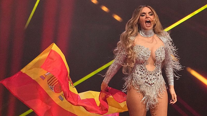 Cantante española en escenario televisivo, con body brillante y medias de rejilla, sosteniendo una bandera de España.  Expresión enérgica.