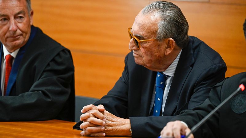 Carlos Fabra, con traje oscuro y gafas de sol, sentado en una mesa de madera durante una vista judicial.  A su lado, un miembro del tribunal.
