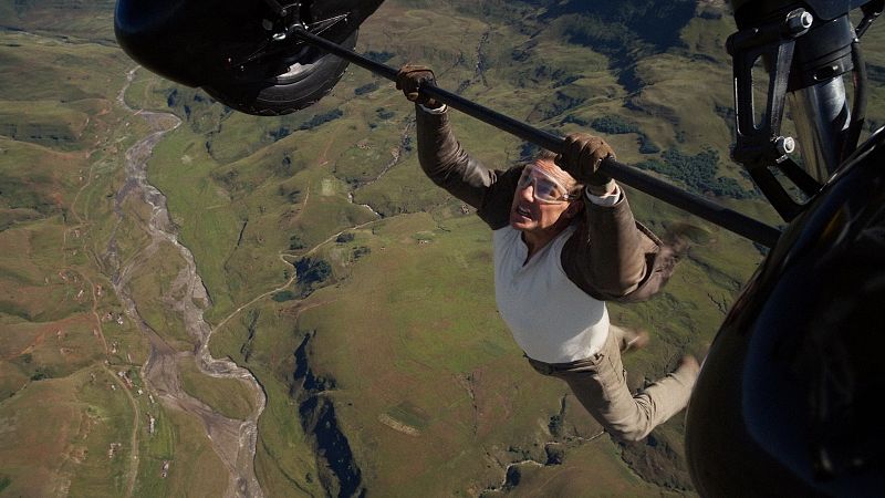 Actor colgado de una barra a gran altura, con gafas protectoras, camiseta blanca y pantalones beige, sobre un paisaje montañoso.