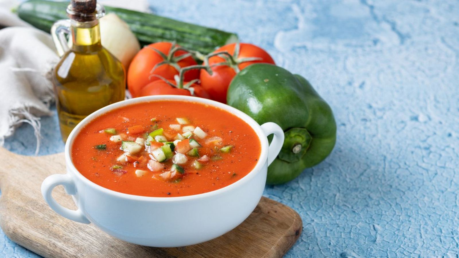 Gazpacho en un tazón blanco con asas, rodeado de sus ingredientes frescos sobre una tabla de madera.  Texto "Gazpacho" y "Noticias de cocina" en la imagen.