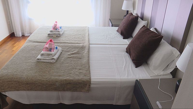 Alojamiento turístico con cama, sábanas blancas, manta beige, mesitas de noche, lámparas, toallas y artículos de aseo. Suelo de madera y ventana con cortina.