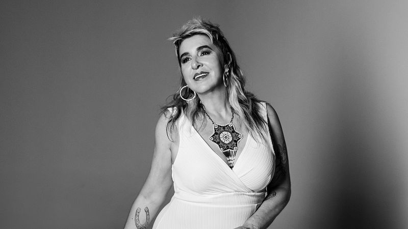 Retrato en blanco y negro de una mujer con vestido blanco, collar y pendientes, mostrando un tatuaje en el brazo y una expresión serena.