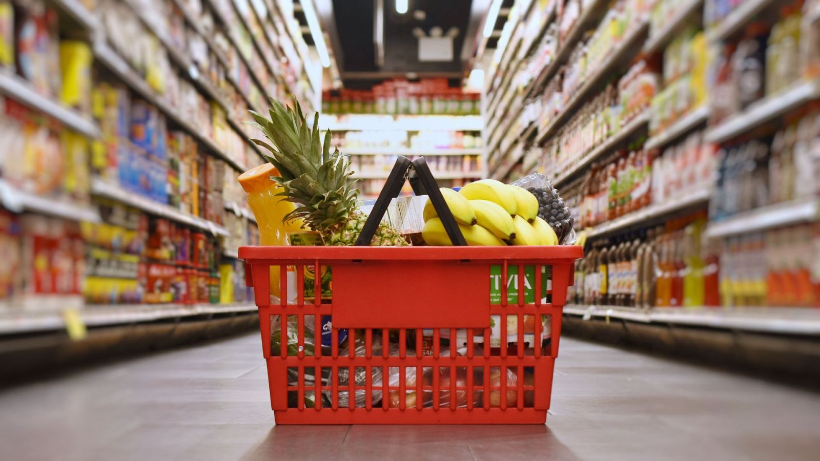 Cesta roja de compra en pasillo de supermercado con plátanos, piña y zumo de naranja visibles.  Estanterías llenas de productos a ambos lados.