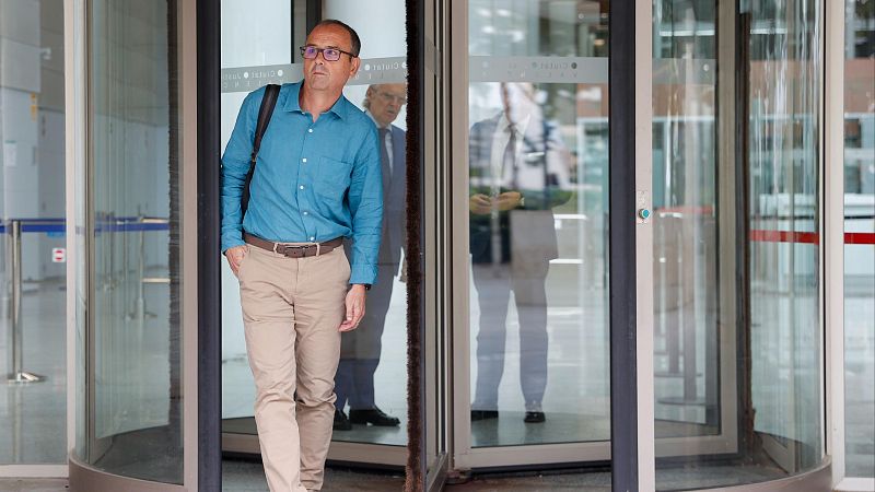Hombre de mediana edad con camisa azul y pantalones beige sale de un edificio a través de una puerta giratoria, llevando una mochila. 