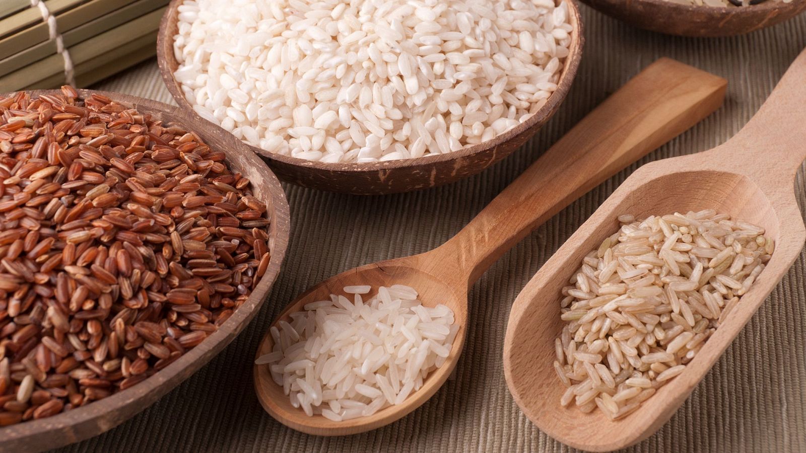 Variedad de arroces en tres cuencos y dos utensilios de madera: arroz integral rojizo, arroz blanco de grano corto y arroz beige claro.  Fondo beige texturizado.