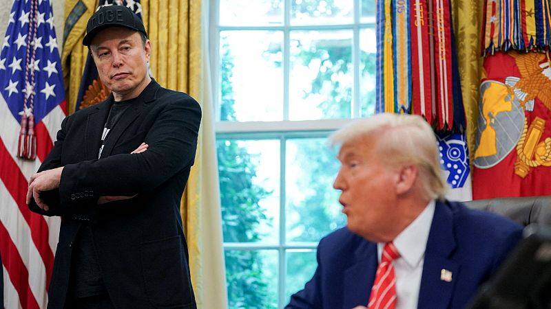 Reunión entre Elon Musk, con gorra negra, y Donald Trump, con traje azul marino y corbata roja.  El escenario parece una oficina con banderas estadounidenses y militares.