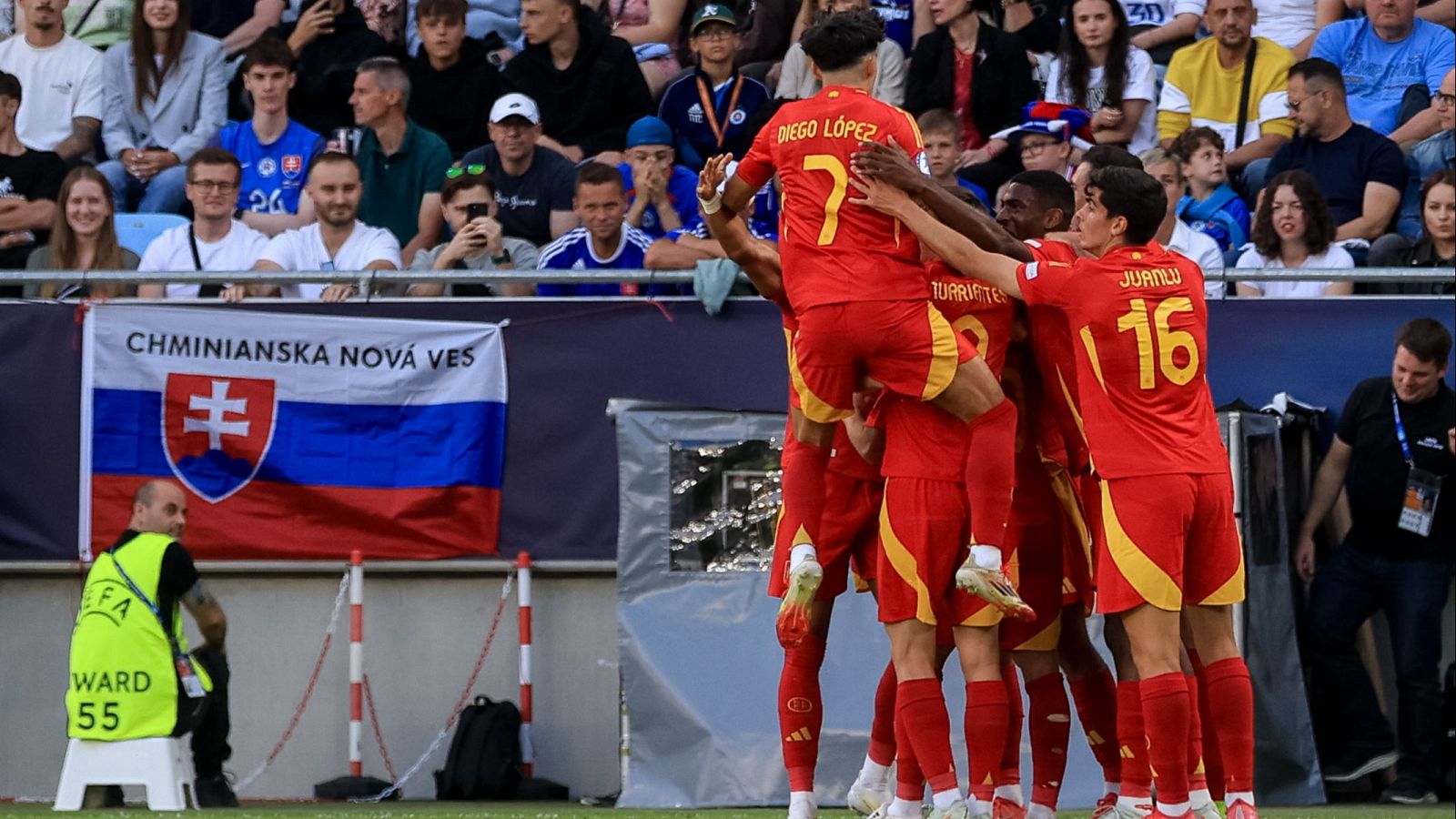 Celebración de gol de la selección española sub-21 cerca de la portería. Se aprecian dorsales de jugadores y una pancarta en el fondo.  Aficionados en las gradas.