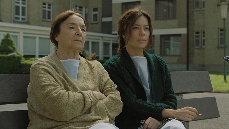 Dos mujeres, una mayor y otra joven con batas, sentadas en un banco exterior observan algo fuera de la imagen.  La escena parece un fotograma de una película.