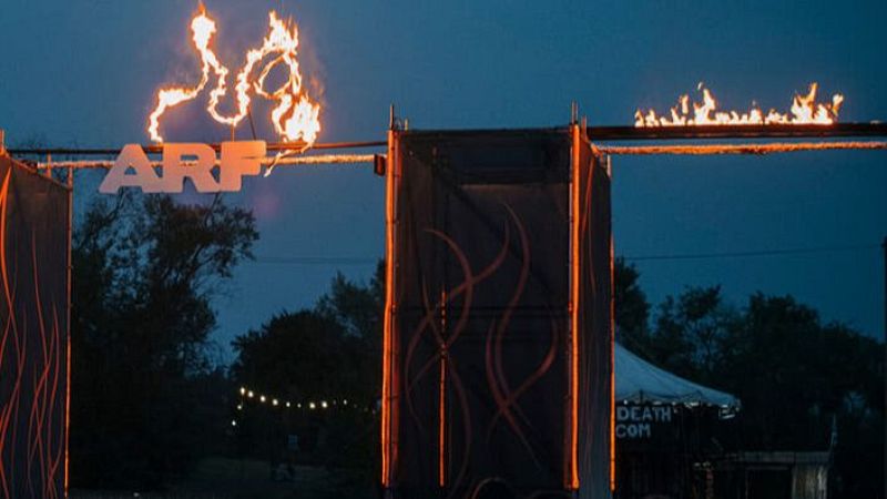 Letras "ARF" en llamas, iluminando la entrada nocturna de un festival. Estructuras con diseños de fuego a los lados, árboles y luces en el fondo.