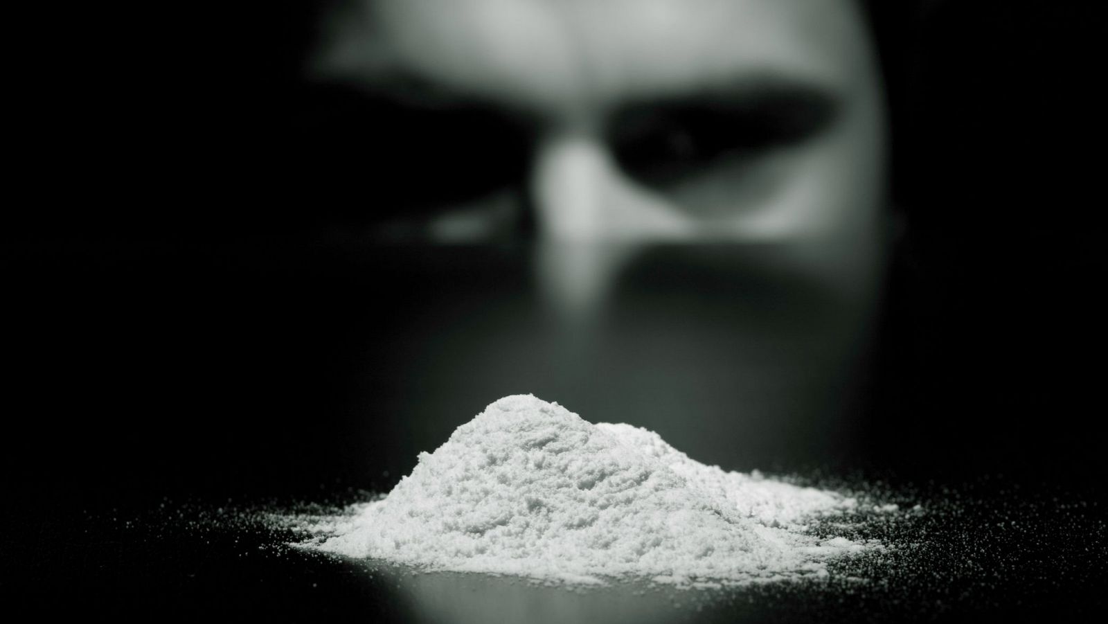 Imagen en blanco y negro de un cúmulo de polvo blanco; una cara observa la sustancia desde un fondo borroso.  Sugiere el consumo de drogas.