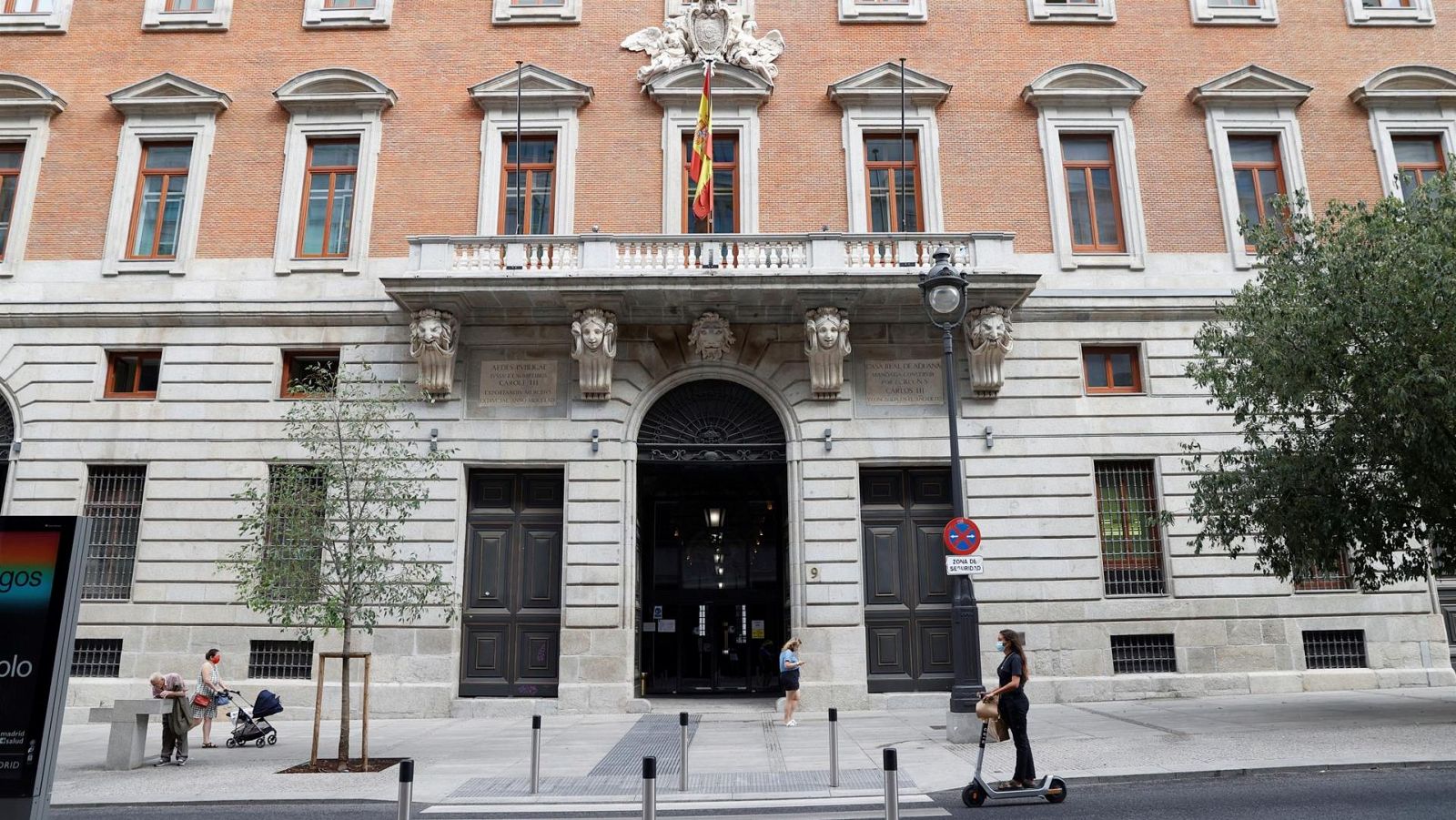 Fachada del Ministerio de Hacienda español con bandera nacional, personas en la acera (mujer con carrito, otra en patinete, persona mayor) y detalles arquitectónicos. 