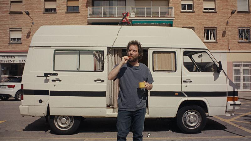 Un hombre con barba y pelo rizado cepilla sus dientes junto a una furgoneta blanca aparcada;  la puerta lateral está abierta y se ve una antena en el techo.