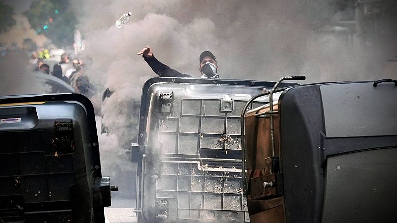 Hombre lanzando botella en una protesta con humo y contenedores volcados como barricadas.  Tensión y confrontación.