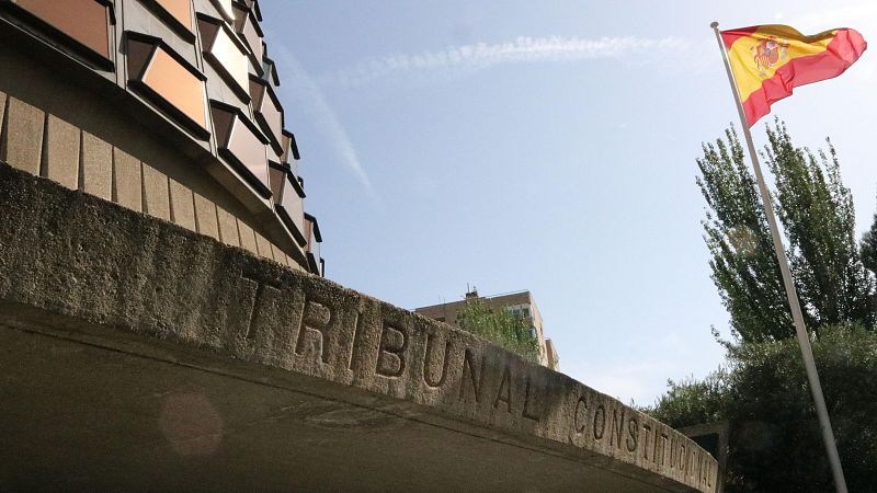 Fachada del Tribunal Constitucional con inscripción en piedra, paneles dorados superiores y bandera española. Cielo despejado.