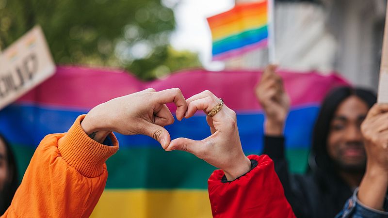Manos formando un corazón con mangas naranja y roja, fondo con banderas arcoíris y bisexual, y personas sosteniendo una bandera y un cartel con "Proud".