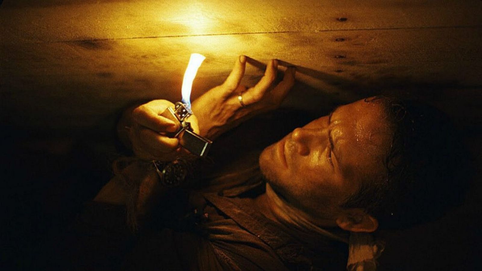 Hombre con mechero en espacio confinado, iluminando su rostro con expresión tensa. Escena de película o serie de televisión estrenada la semana del 7 de julio.
