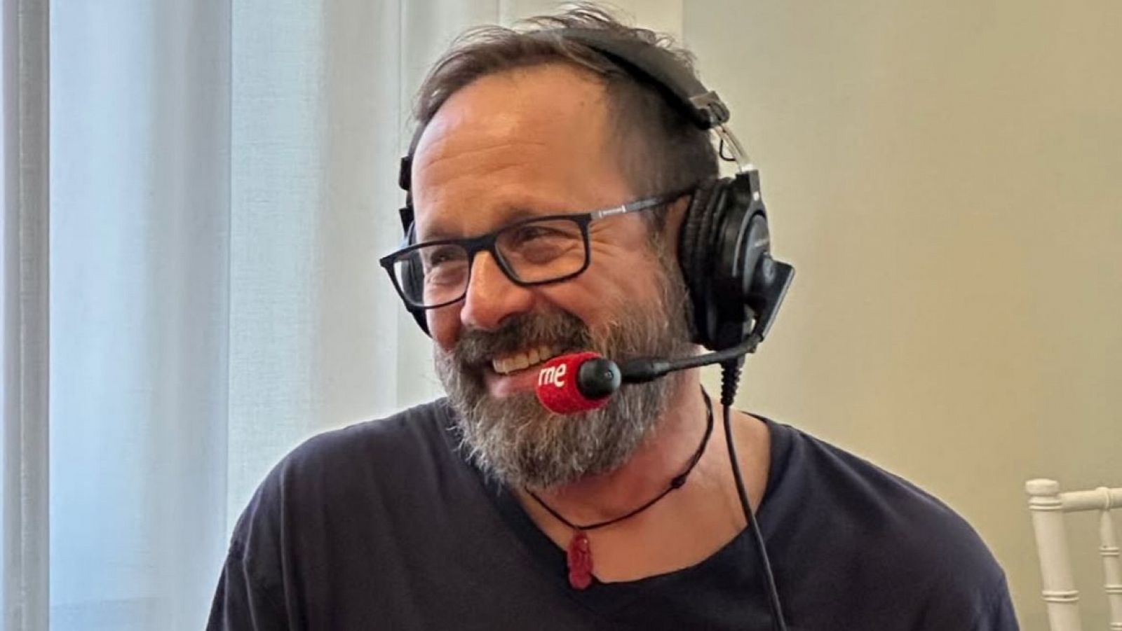 Hombre de mediana edad con barba, gafas y auriculares, sonriendo y usando un micrófono con una esponja roja que dice "me".  Fondo con cortina y silla.
