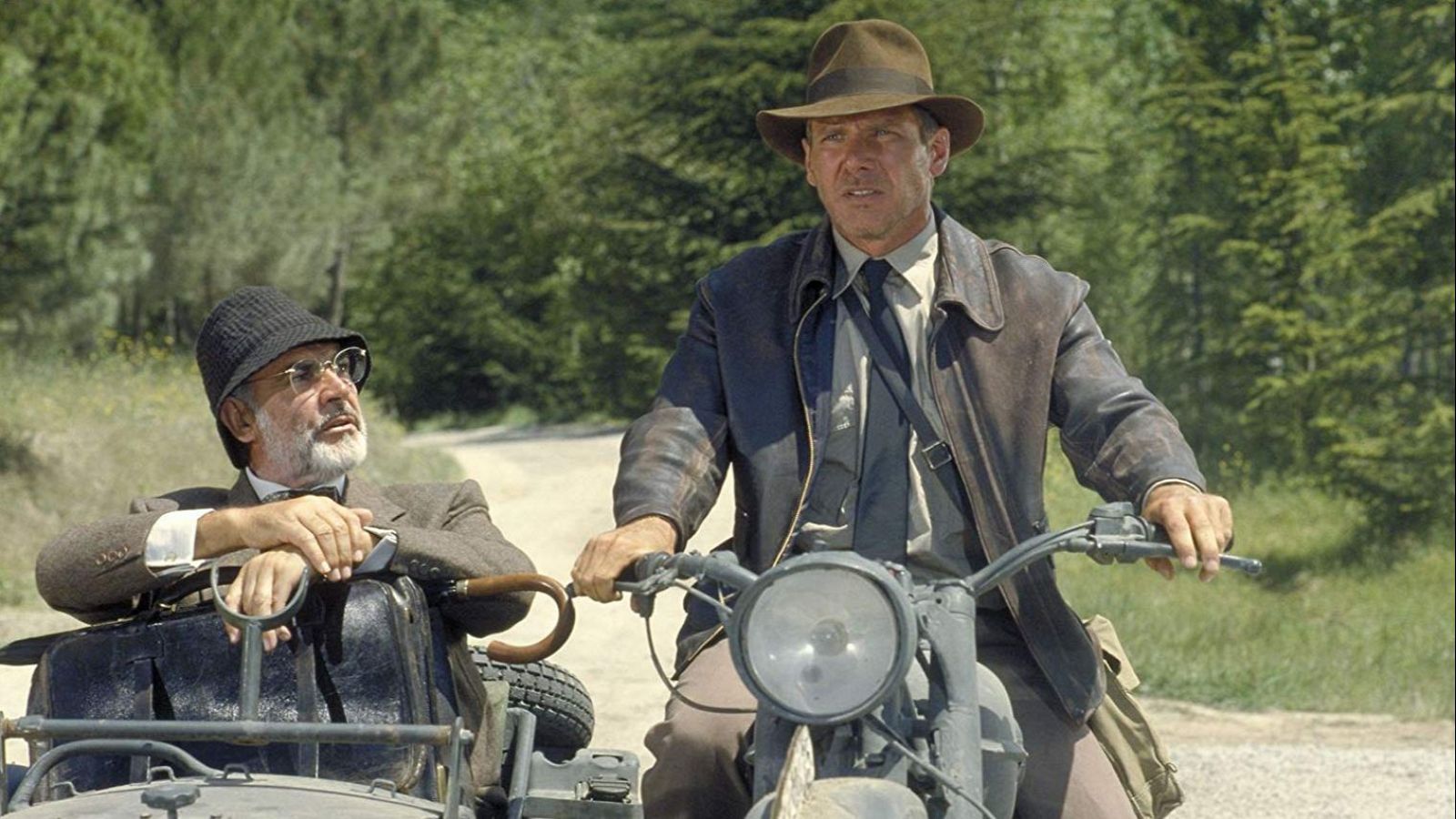 Indiana Jones (Harrison Ford) en motocicleta con sidecar, donde viaja acompañado por un hombre con traje y bastón (Sean Connery). Escena en camino de tierra arbolado.
