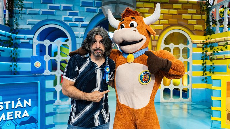 Humorista con camisa estampada y vaqueros señala a una vaca marrón anaranjada con traje azul, mascota de un programa de televisión, que hace un gesto de 'pulgar arriba'.