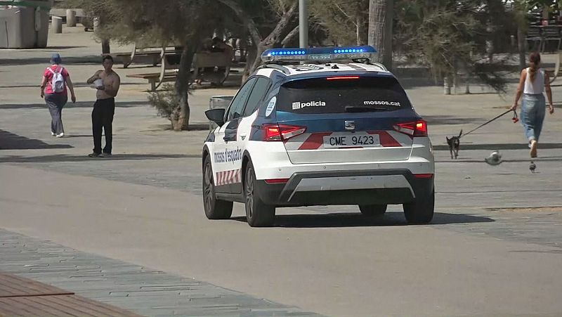 Coche patrulla Mossos d'Esquadra (matrícula CME 9423) con luces azules, circulando por zona peatonal con personas y vegetación.  'mossos.cat' y 'policia' visibles en el vehículo.