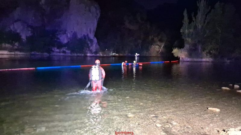 Búsqueda nocturna de desaparecidos en río. Bomberos con trajes de buceo trabajan en aguas poco profundas usando una barrera flotante. Escena dramática.