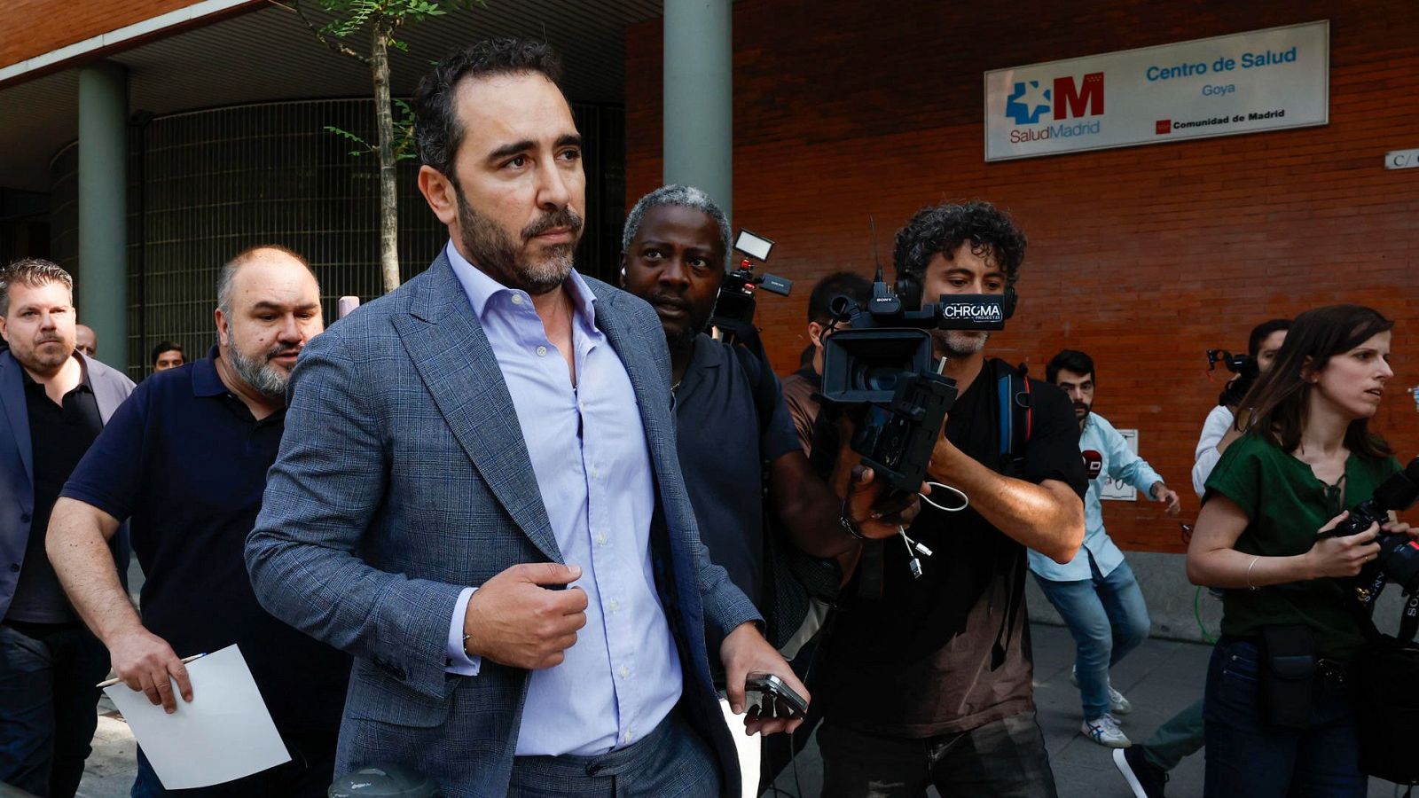 Un hombre en americana gris y camisa azul sale de un centro de salud rodeado de periodistas con cámaras y micrófonos.