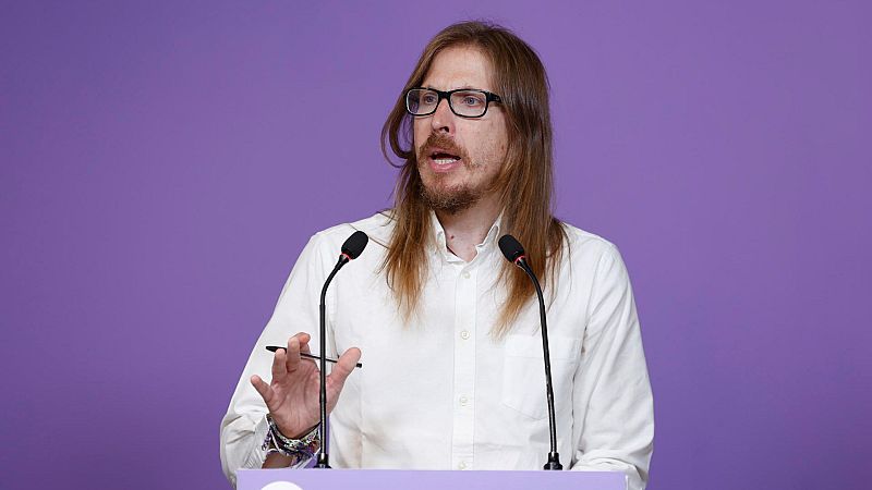 Portavoz con camisa blanca y gafas ofrece rueda de prensa tras atril morado con dos micrófonos; sostiene bolígrafo.