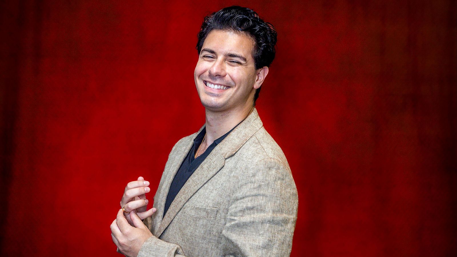 Fotografía de Alfred García sonriendo, con americana beige y camiseta oscura, manos entrelazadas sobre fondo rojo.  Expresión relajada y positiva.