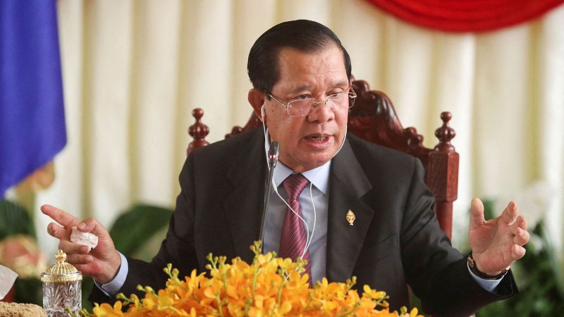 Rueda de prensa: Primer ministro sentado, traje oscuro, corbata granate, gesticulando.  Sobre la mesa, flores y un recipiente de cristal.