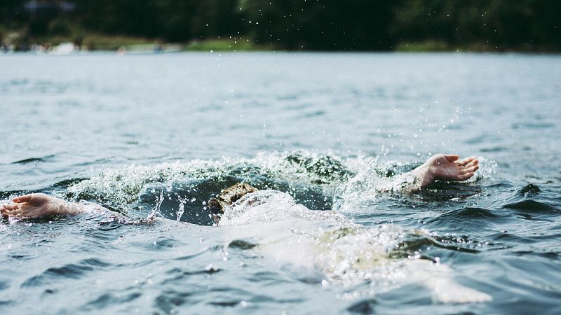 Imagen de una persona aparentemente ahogándose en el mar; cuerpo boca abajo, brazos extendidos.  El agua es oscura y tranquila, con pequeñas ondas.