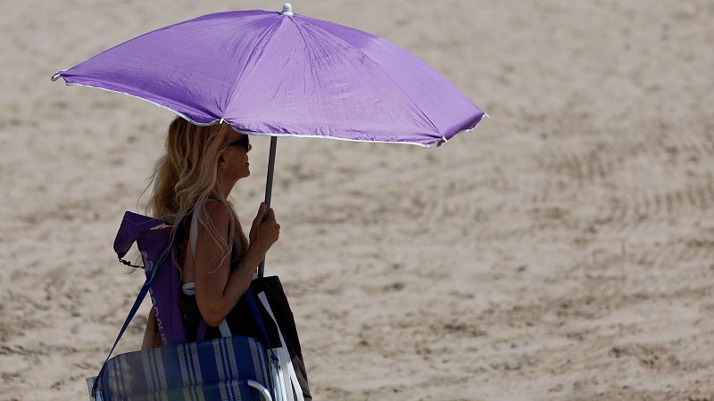 Mujer en la playa con sombrilla morada, gafas de sol y bikini, protegiéndose del sol con una silla plegable y una bolsa de playa.