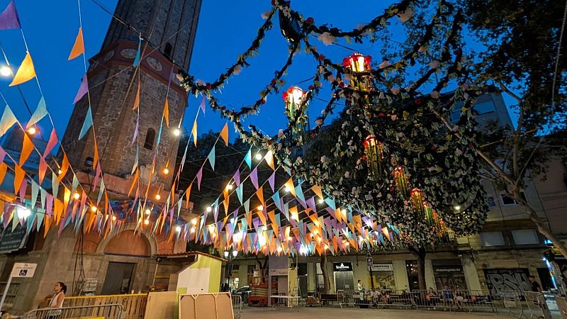 Plaza decorada festivamente al anochecer.  Se ven guirnaldas, banderines y luces. Personas disfrutando en mesas.