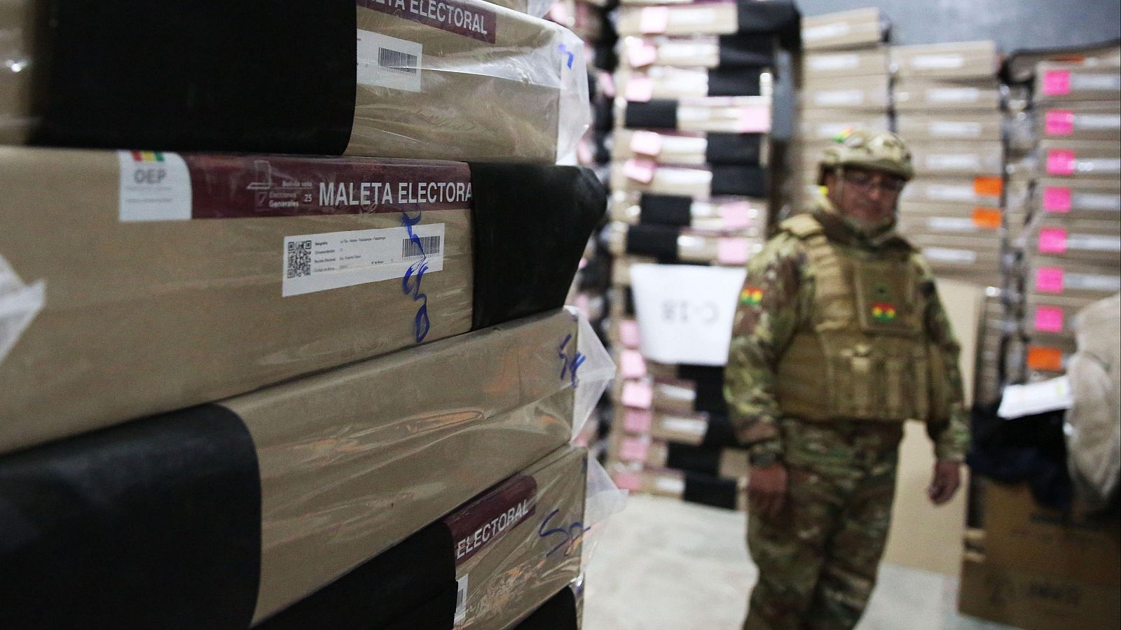 Maletas electorales de cartón, con códigos QR y marcas azules, son custodiadas por un militar boliviano con uniforme de camuflaje y la bandera nacional en su equipo.