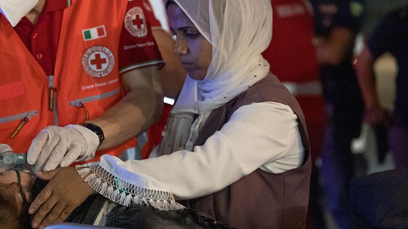 Trabajador humanitario de la Cruz Roja Italiana asiste a una persona con máscara de oxígeno.  Una mujer con hiyab observa cerca.  Escena de emergencia médica.