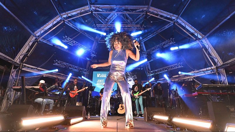 Actuación musical en directo con cantante en mono plateado, iluminada por focos azules y blancos.  Se observa el logo de Radio 3 en el escenario.