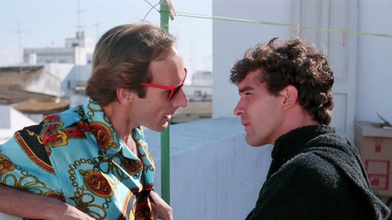 Escena de película con Eusebio Poncela, camisa estampada y gafas rojas, mirando a Antonio Banderas, quien viste ropa oscura. Fondo con tendedero.