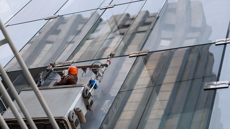 Trabajador en plataforma elevadora limpiando fachada de cristal de un edificio alto. Lleva casco naranja y ropa de trabajo.