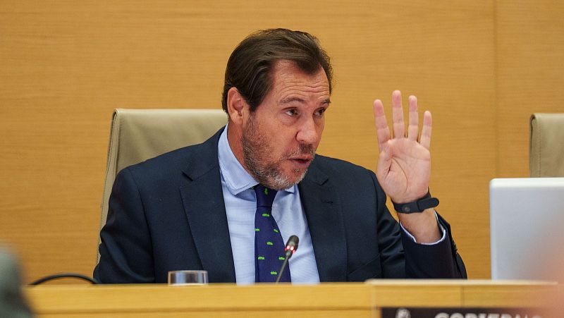 Ministra en reunión, traje azul oscuro, gesticulando mientras habla.  Sobre la mesa, micrófono y vaso de agua. Fondo de madera clara.