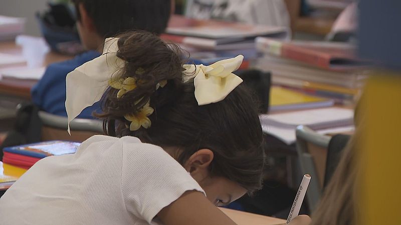 Niña sentada en pupitre escolar escribiendo en un cuaderno, con el pelo recogido y una camiseta blanca. Fondo desenfocado con otros alumnos y materiales.