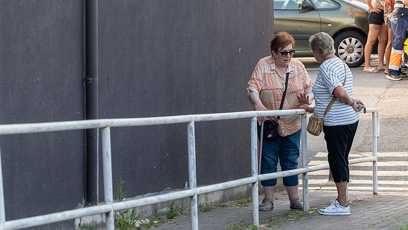 Dos mujeres mayores conversan junto a una barandilla. Una lleva blusa estampada y vaqueros; la otra, camiseta a rayas y pantalones negros.  Fondo urbano con coche visible.