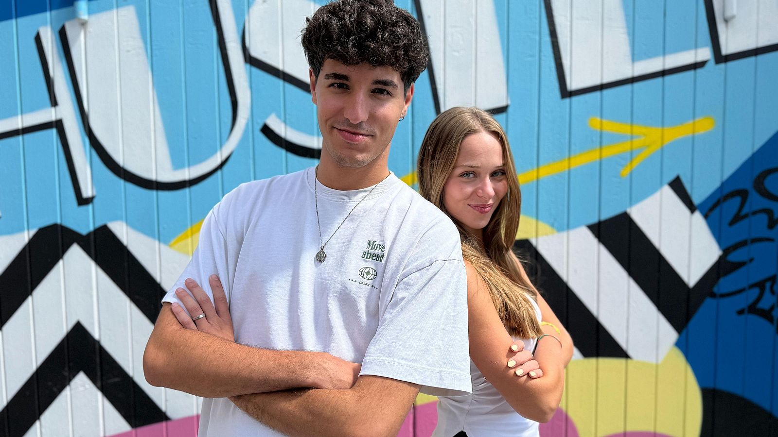 Jóvenes con camisetas blancas posan ante un mural multicolor con formas geométricas. Él lleva un collar; ella, el pelo largo. Mirada relajada a cámara.