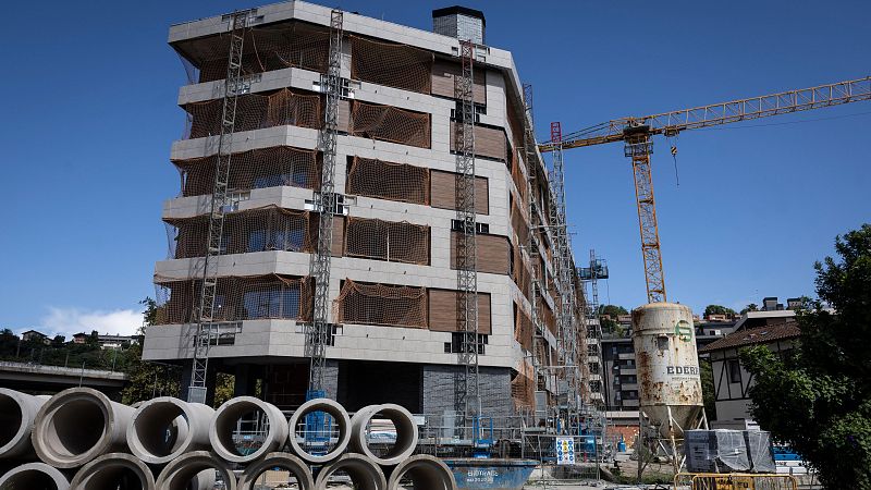 Obra en progreso: edificio multiplanta con andamios, grúa, tuberías y contenedor de escombros. Fachada gris y marrón. Cielo despejado.