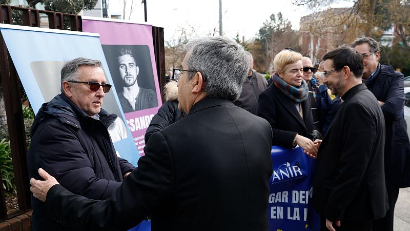 El presidente y vicepresidente de la Conferencia Episcopal Española saludan a víctimas de abusos sexuales en un acto público.  Se aprecian carteles con imágenes y texto fragmentado en el fondo.