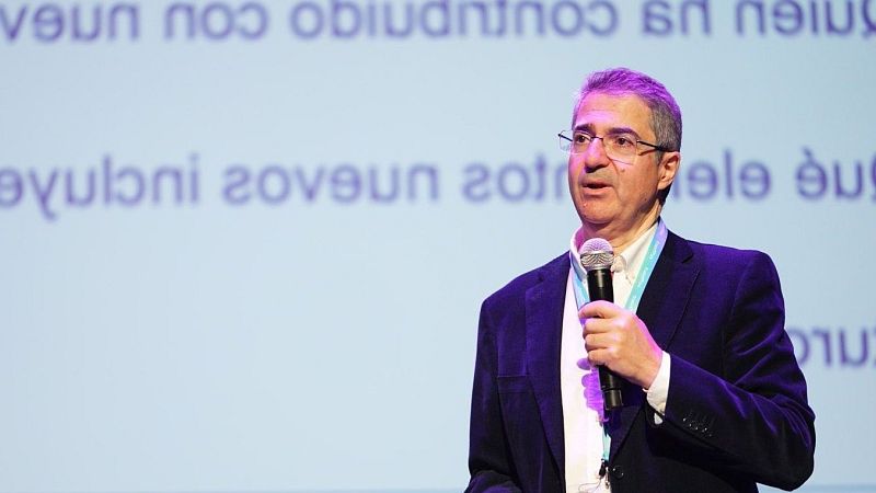 Catedrático de la UPM dando una presentación con micrófono, traje oscuro y camisa blanca. Fondo con pantalla de proyección y texto indistinguible.