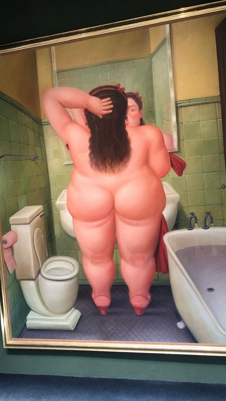 Una obra de arte muestra a una mujer de espaldas, con una figura voluminosa, mirándose en un espejo dentro de un baño con azulejos verdes. La mujer tiene el pelo oscuro recogido con una cinta roja.