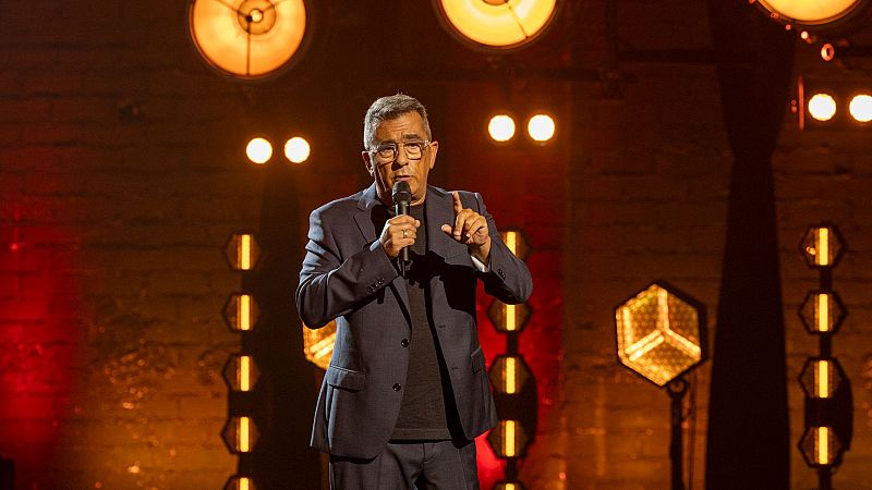 En un escenario iluminado, Andreu Buenafuente, con traje oscuro y camiseta negra, sostiene un micrófono y gesticula con el dedo índice, posiblemente durante una intervención.
