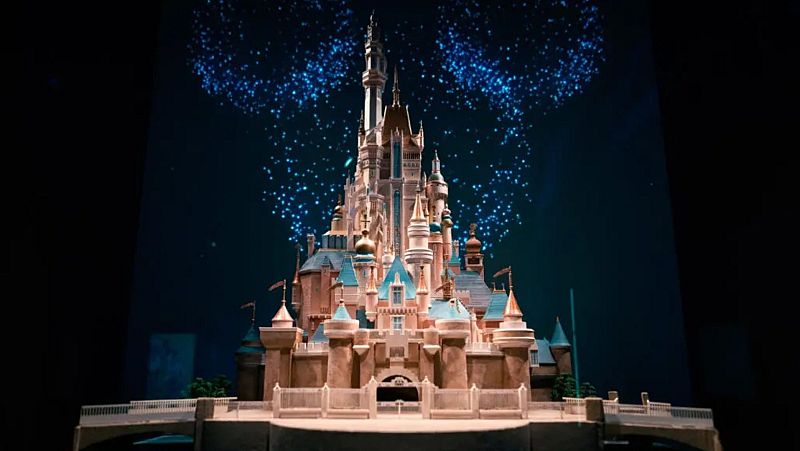 Una maqueta iluminada de un castillo con múltiples torres y detalles arquitectónicos, con colores beige, azul y dorado, y un fondo oscuro con fuegos artificiales azules.