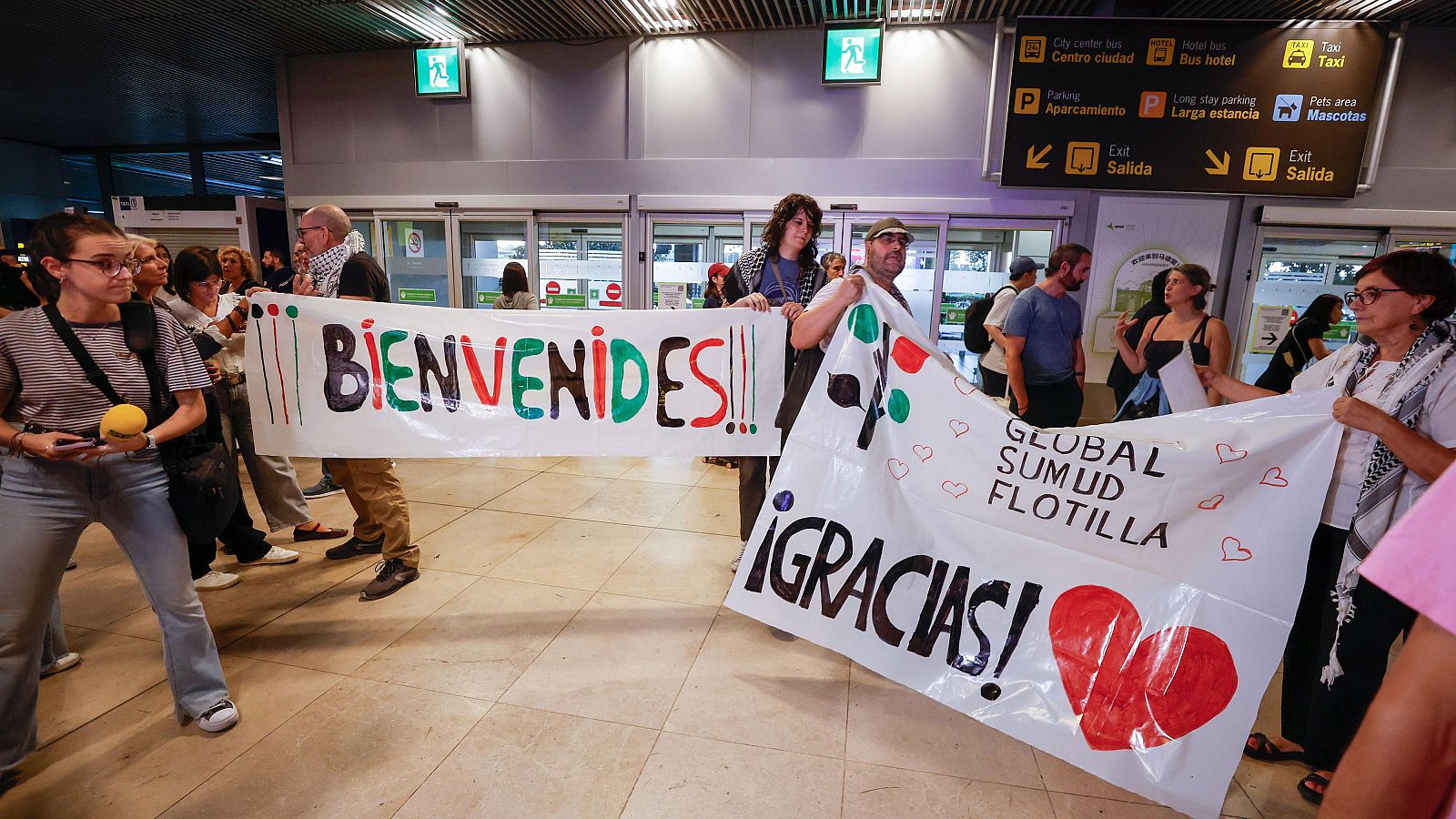 Aterriza en Madrid el avión con 21 activistas españoles de la flotilla retenidos en Israel En la zona de llegadas de un aeropuerto, un grupo de personas sostiene pancartas de bienvenida y agradecimiento. Se observan carteles con mensajes coloridos y corazones, en un ambiente de celebración.